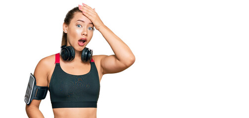 Beautiful blonde woman wearing gym clothes and using headphones surprised with hand on head for mistake, remember error. forgot, bad memory concept.