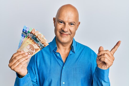 Middle age bald man holding canadian dollars smiling happy pointing with hand and finger to the side