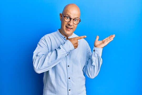 Middle age bald man wearing casual clothes and glasses amazed and smiling to the camera while presenting with hand and pointing with finger.