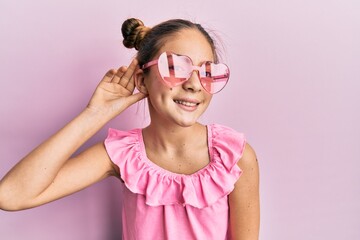 Beautiful brunette little girl wearing heart shaped sunglasses smiling with hand over ear listening and hearing to rumor or gossip. deafness concept.