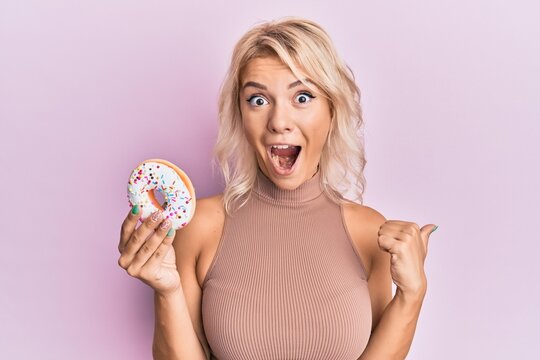 Young blonde girl holding donut pointing thumb up to the side smiling happy with open mouth