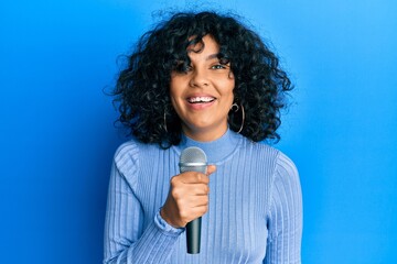 Young hispanic woman singing song using microphone looking positive and happy standing and smiling with a confident smile showing teeth