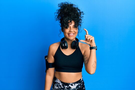 Young hispanic woman wearing gym clothes and using headphones smiling and confident gesturing with hand doing small size sign with fingers looking and the camera. measure concept.