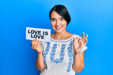 Beautiful young woman with short hair holding paper with love is love message doing ok sign with fingers, smiling friendly gesturing excellent symbol