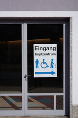 Sign on glass door saying: Vaccination center entrance (germany: Eingang Impfzentrum). Bright exterior facade, the surroundings reflected in the glass. Front view.
