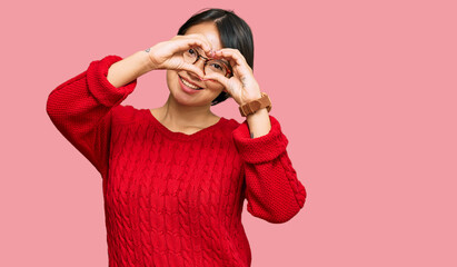 Young beautiful hispanic woman with short hair wearing casual sweater and glasses doing heart shape with hand and fingers smiling looking through sign