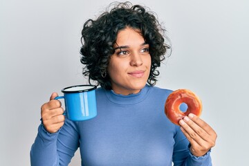 Young hispanic woman with curly hair drinking a cup of coffee and croissant smiling looking to the side and staring away thinking.