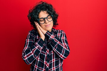Fototapeta na wymiar Young hispanic woman with curly hair wearing casual clothes and glasses sleeping tired dreaming and posing with hands together while smiling with closed eyes.