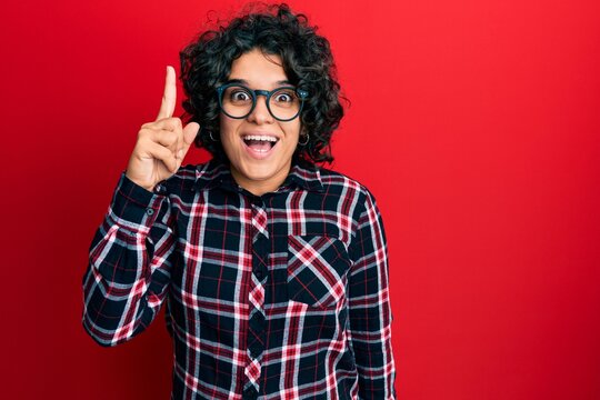 Young hispanic woman with curly hair wearing casual clothes and glasses pointing finger up with successful idea. exited and happy. number one.