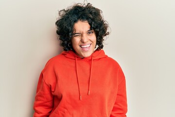 Young hispanic woman with curly hair wearing casual sweatshirt winking looking at the camera with...