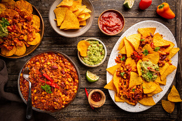 Mexican food concept: tortilla chips, guacamole, salsa, chilli con carne and fresh ingredients over wooden background, top view