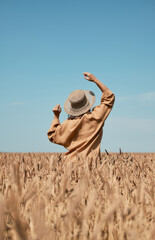 Happy girl in a hat dancing in a field on a clear blue sky background. Hands are raised up. Back view.
