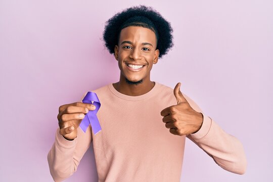 African american man with afro hair holding awareness purple ribbon smiling happy and positive, thumb up doing excellent and approval sign