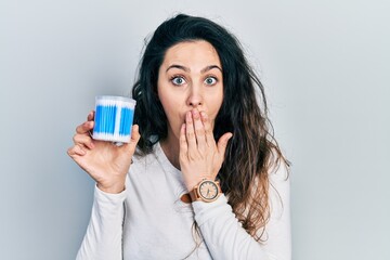 Young hispanic woman holding earwax cotton remover covering mouth with hand, shocked and afraid for mistake. surprised expression