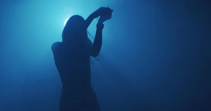 Dancing young girl in cloud of smoke, blue night club light. Modern dance, 4k video. Mysterious moving female silhouette