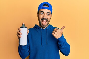 Young hispanic man wearing sweatshirt holding graffiti spray pointing thumb up to the side smiling happy with open mouth