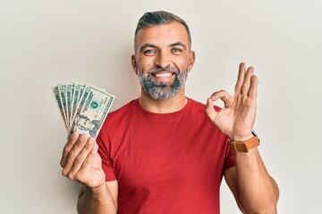 Middle age handsome man holding 20 dollars banknotes doing ok sign with fingers, smiling friendly gesturing excellent symbol