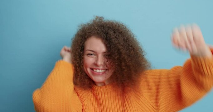 Cheerful overjoyed curly haired millennial girl jumps from joy dances carefree expresses positive emotions wears casual orange sweater isolated over blue background. People and amusement concept
