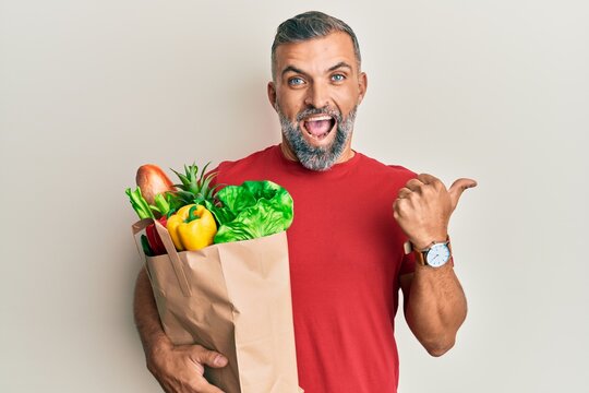 Middle age handsome man holding paper bag with bread and groceries pointing thumb up to the side smiling happy with open mouth