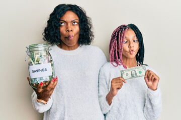 Beautiful african american mother and daughter holding jar with savings and 1 dollar banknote making fish face with mouth and squinting eyes, crazy and comical.