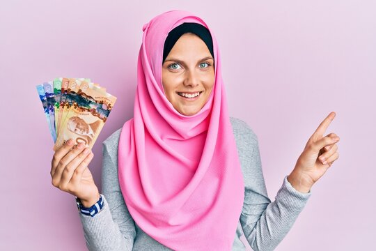 Young caucasian woman wearing islamic hijab holding canadian dollars banknotes smiling happy pointing with hand and finger to the side