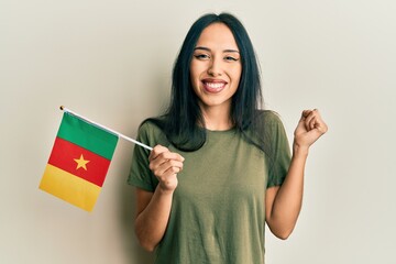 Young hispanic girl holding cameroon flag screaming proud, celebrating victory and success very...