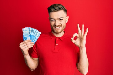 Young redhead man holding 10000 chilean pesos doing ok sign with fingers, smiling friendly gesturing excellent symbol