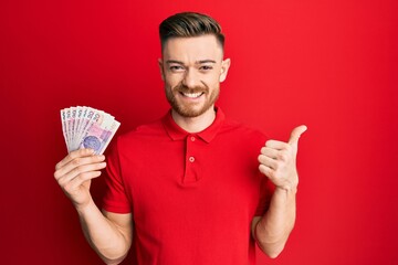 Young redhead man holding 20 polish zloty banknotes smiling happy and positive, thumb up doing excellent and approval sign