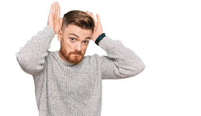 Fototapeta premium Young redhead man wearing casual winter sweater doing bunny ears gesture with hands palms looking cynical and skeptical. easter rabbit concept.