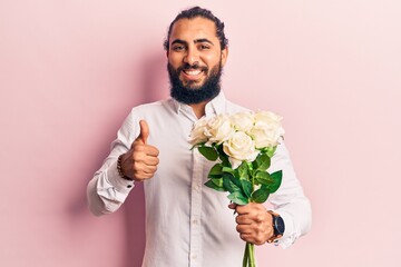 Young arab man holding flowers smiling happy and positive, thumb up doing excellent and approval sign