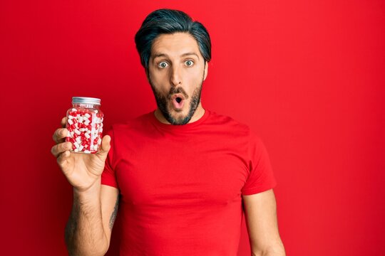 Young hispanic man holding jar of pills scared and amazed with open mouth for surprise, disbelief face