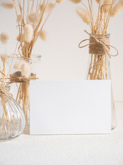 Blank white greeting card mock up.decoration with dried Lagurus ovatus flowers composition in modern glass vase  on  beige table and cement wall background