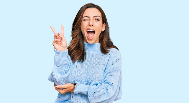 Young brunette woman wearing casual winter sweater smiling with happy face winking at the camera doing victory sign with fingers. number two.