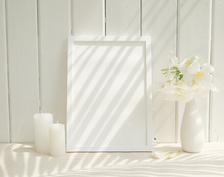 Blank mockup picture frame,white candle,and Plumeria flower in ceramic white vase on beige table with white wood background,long palm shadow