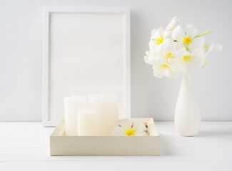 White candles on white wood  tray,mock up poster frame and Plumeria flowers in modern ceramic vase  over white wooden table room interior,spa soft tone still life
