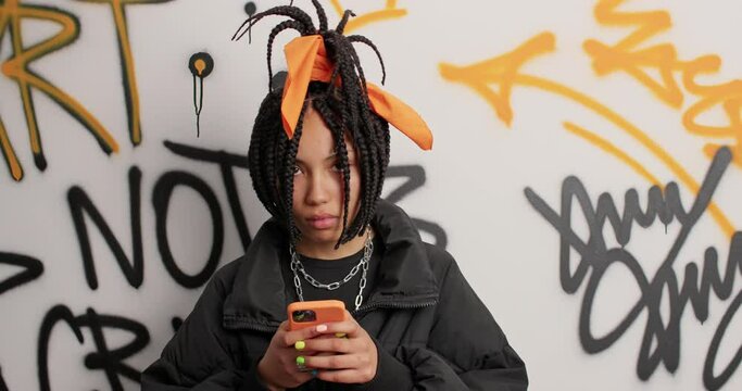 Trendy hipster girl chews bubble gum types messages via smartphone wears black jacket has braid hairstyle being addicted to modern technologies poses against graffiti wall. Teen lifestyle concept