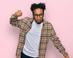 Young african american man with beard wearing casual clothes and glasses angry and mad raising fist frustrated and furious while shouting with anger. rage and aggressive concept.