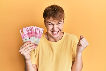 Young caucasian man holding 100000 indonesian rupiah screaming proud, celebrating victory and success very excited with raised arm