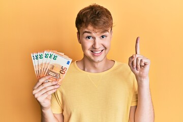 Young caucasian man holding bunch of 50 euro banknotes smiling with an idea or question pointing...