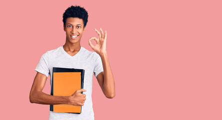 Young african american man holding book doing ok sign with fingers, smiling friendly gesturing excellent symbol
