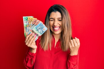 Beautiful brunette woman holding australian dollars very happy and excited doing winner gesture with arms raised, smiling and screaming for success. celebration concept.