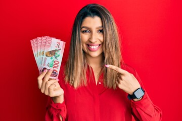 Beautiful brunette woman holding 100 new zealand dollars banknote smiling happy pointing with hand and finger