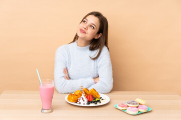 Teenager girl eating waffles isolated on beige background looking up while smiling