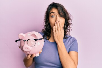 Young hispanic woman holding piggy bank with glasses covering mouth with hand, shocked and afraid...