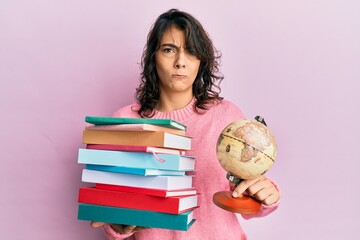 Young hispanic woman holding a pile of books and world ball skeptic and nervous, frowning upset...