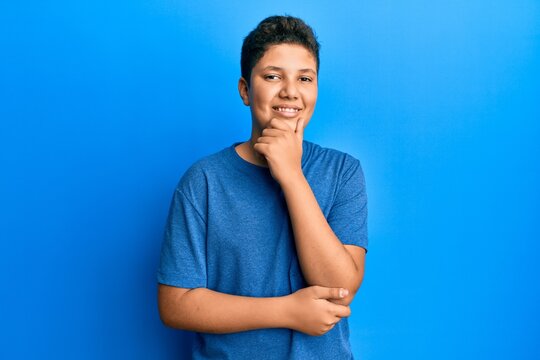 Teenager hispanic boy wearing casual blue t shirt smiling looking confident at the camera with crossed arms and hand on chin. thinking positive.