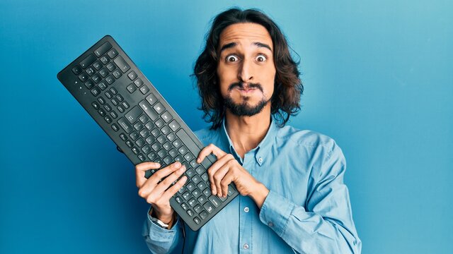Young hispanic man holding keyboard puffing cheeks with funny face. mouth inflated with air, catching air.