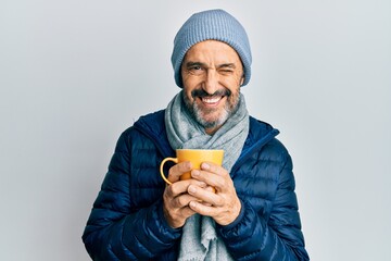 Middle age hispanic man wearing winter scarf and drinking a cup of hot coffee winking looking at...