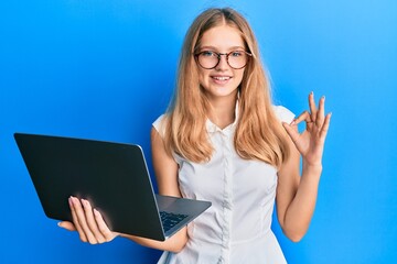 Beautiful young caucasian girl working using computer laptop doing ok sign with fingers, smiling friendly gesturing excellent symbol