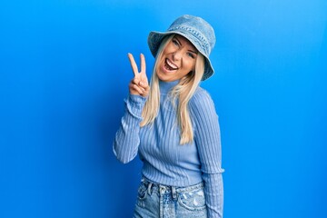 Obraz na płótnie Canvas Young blonde woman wearing casual denim hat smiling with happy face winking at the camera doing victory sign. number two.